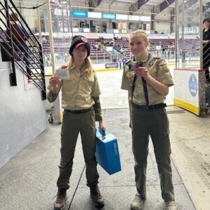 Two Boy Scouts in uniform holding up gift cards in front of the ice at a hockey arena